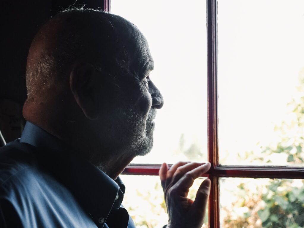 An elderly man gazing out of a sunlit window, conveying a sense of reflection and solitude. The image highlights themes of emotional resilience and strategies to cope with loneliness and isolation in later life