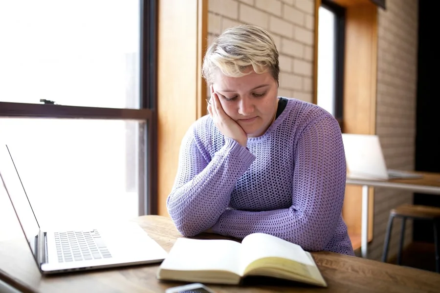 blonde girl reading book next to laptop