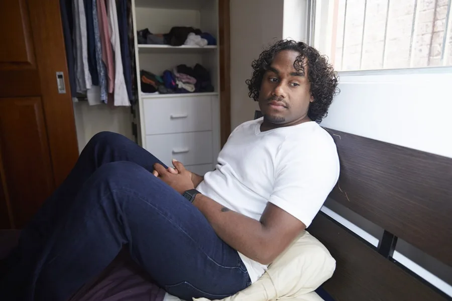 Photo of a boy sitting in his bedroom on his bed with knees up and hands together in his lap. He looks a little overwhelmed and tired.