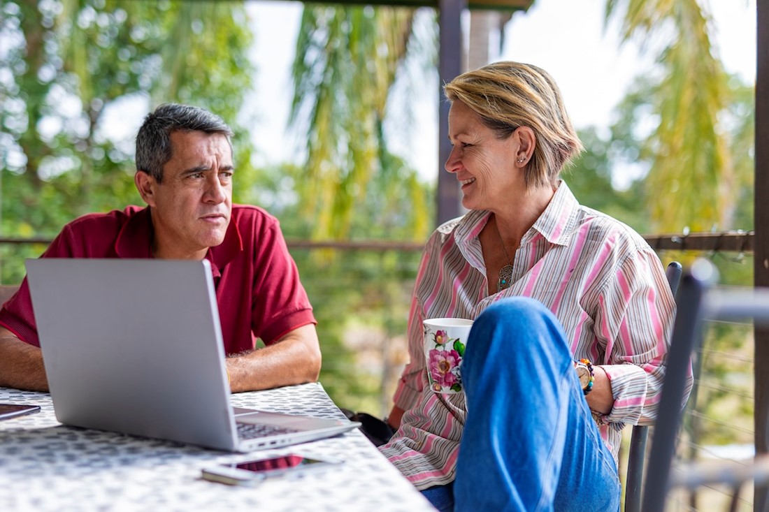 Husband and wife sharing a cup of tea outside on their balcony, discussing something on their laptop.