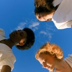 An African woman and two Caucasian women join hands while dancing in a circle outside