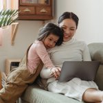 A mother and her young daughter hug while using a laptop together on the couch