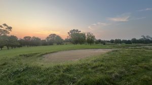 A pink golf ball sits in a sand trap as a group of golfers crests a hill in the distance at twilight and a sunset in the distance
