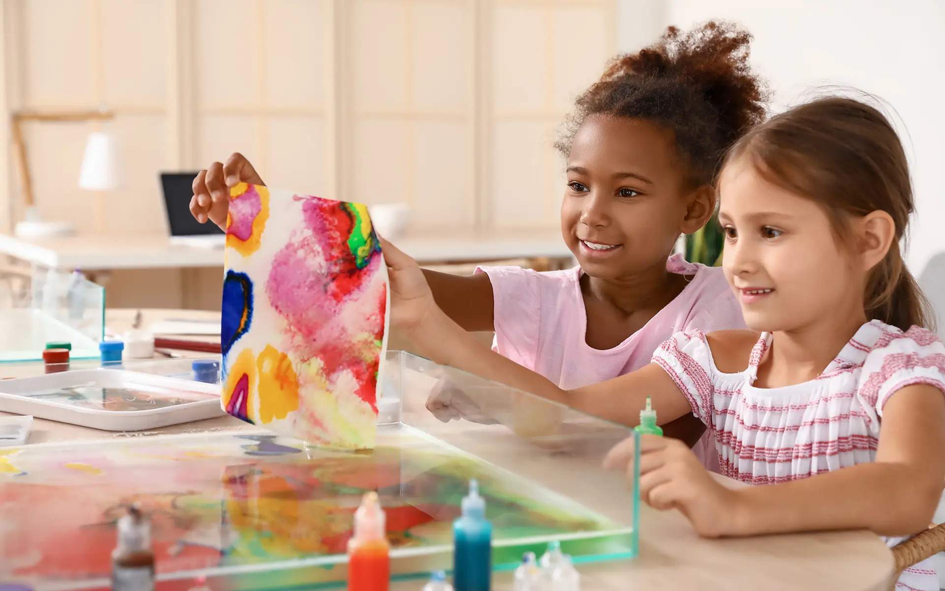 Two young girls smiling while creating colorful marble art with paint and water at a table.
